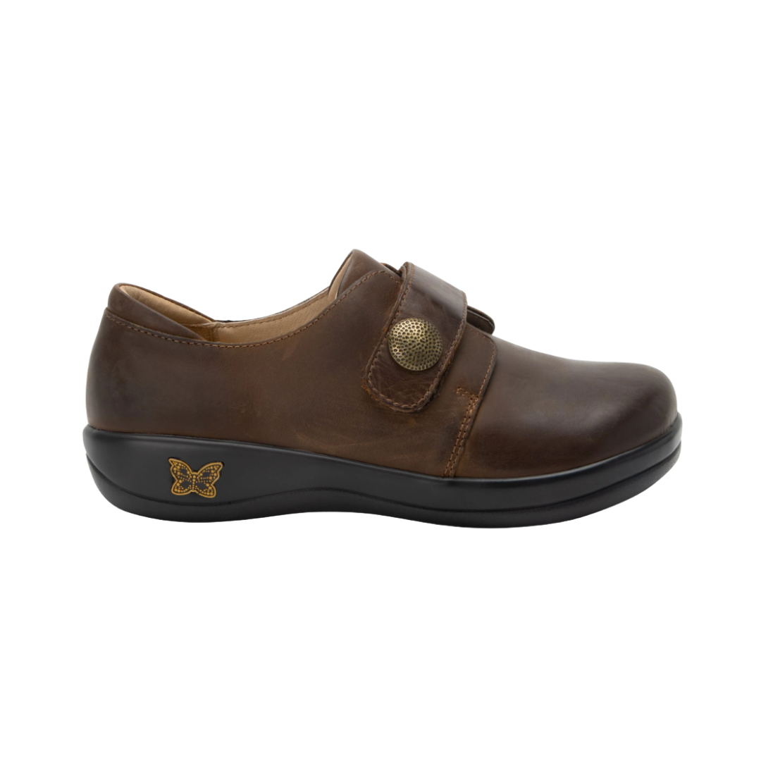 Alegria Women’s Joleen Slip-On Shoe in Oiled Brown side view showcasing the premium leather upper with subtle shine, adjustable hook-and-loop strap, decorative button detail, mild rocker outsole, and professional slip-on design built for comfort and stability.